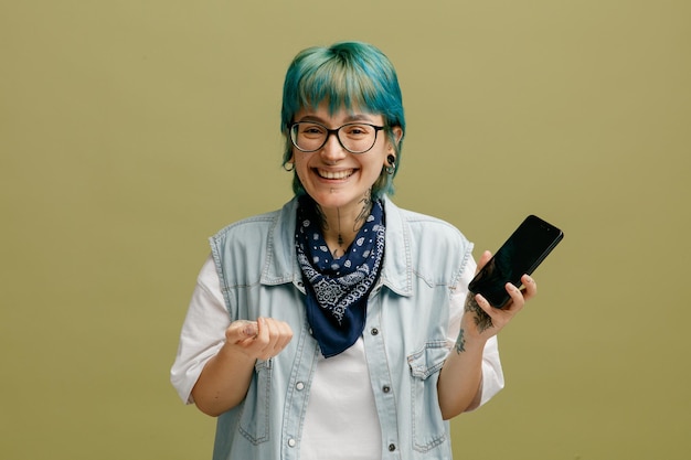 Joyful young woman wearing glasses bandana on neck holding mobile phone looking at camera while keeping hand in air isolated on olive green background