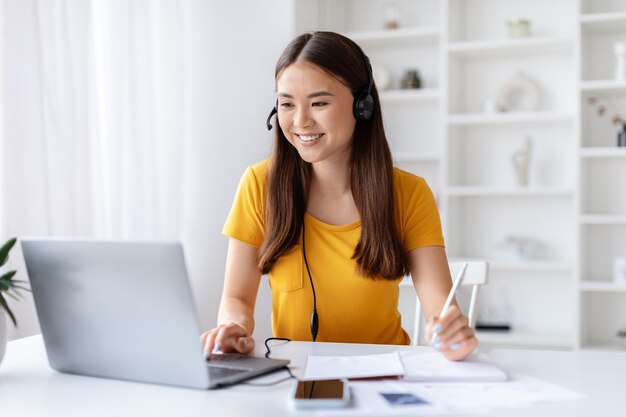 Joyful young woman wearing headset multitasking with pen and laptop at home