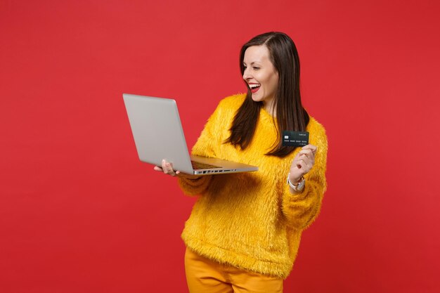 Joyful young woman in yellow fur sweater holding credit bank card, laptop pc computer isolated on bright red wall background in studio. People sincere emotions, lifestyle concept. Mock up copy space.