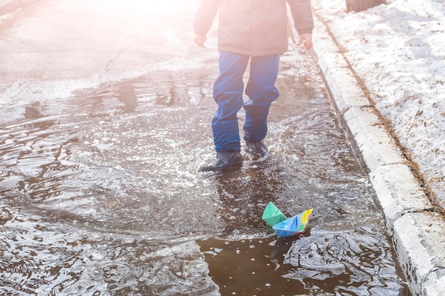 Joyfull Child boy of 67 year old in rubber boots playing with origami paper boats in muddy puddle Carefree childhood happy time