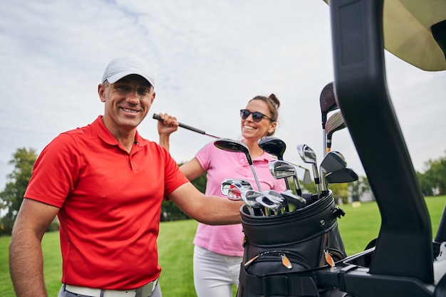 Joyous dark-haired Caucasian female athlete and a professional male trainer standing by a golf car