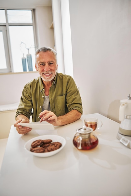 Joyous elderly man with a tablet having tea with biscuits