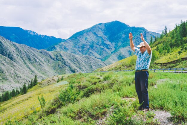 Joyous man near highway among giant mountains. Cheerful traveler on hill among rich vegetation. Mountain tourism. Journey in highland.