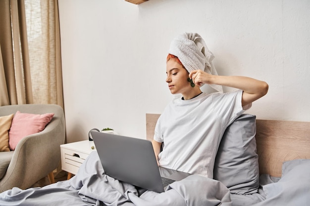 joyous pretty queer person with hair towel watching movies on laptop and using gua sha while in bed