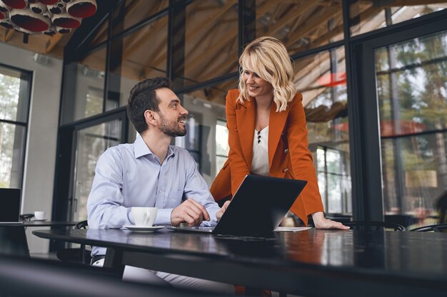 Joyous pretty young woman in a stylish jacket standing next to her Caucasian male coworker