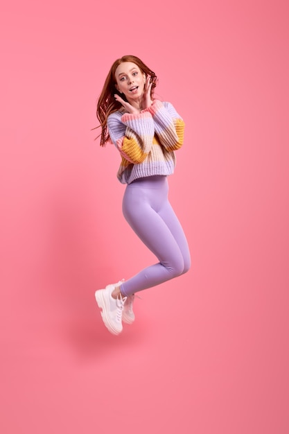 Joyous redhead woman in casual wear have fun alone in studio posing at camera jumping holding hands on cheeks isolated pink studio background portrait