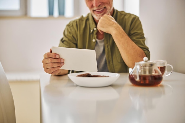 Joyous senior citizen having tea while looking at tablet