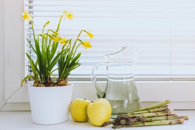 Jug of clean water, apples, asparagus, daffodil flowers, on windowsill, morning, healthy breakfast,