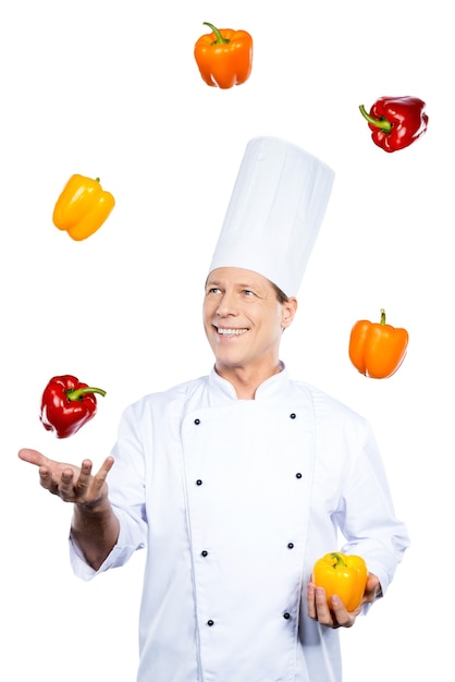 Juggling with colors. Cheerful mature chef in white uniform holding colorful peppers and looking at the red one while standing against white background