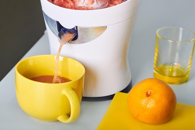 juicer at work freshly squeezed orange juice in yellow cup fresh cut orange on the white table