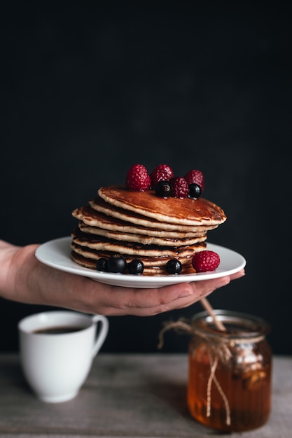 Juicy pancakes with berries and honey on white plate on human hand, jar and spoon, wooden table with cup of coffee. High quality photo