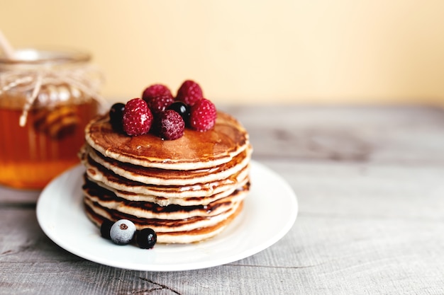 Juicy pancakes with berries and honey on a white plate, spoon, jar, wooden table. High quality photo