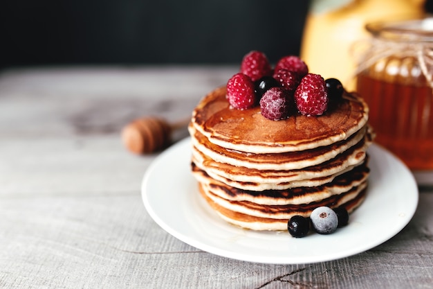 Juicy pancakes with berries and honey on a white plate, spoon, jar, wooden table, yellow vase with eucalyptus. High quality photo