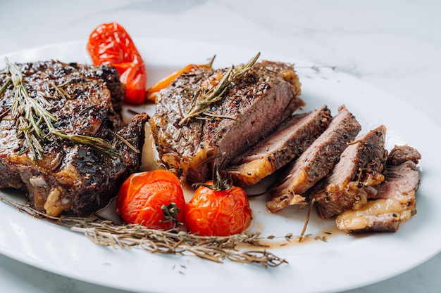 Juicy wellfried ribeye steaks on a white platter on a white marble surface with raspmarine and fried cherry tomatoes and peppers steaks are sliced and ready to serve