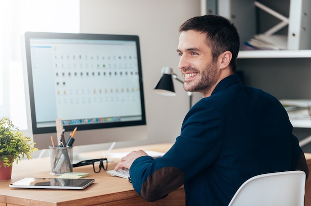 Just inspired. Handsome young man looking away and smiling while sitting at his working place in office