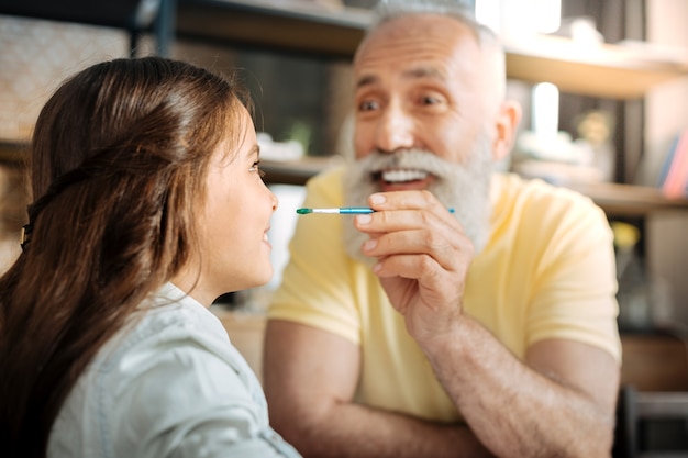 Just kidding. Playful happy senior man holding a brush with a watercolor on it and approaching it to the nose of his little daughter, wanting to put a dot on it