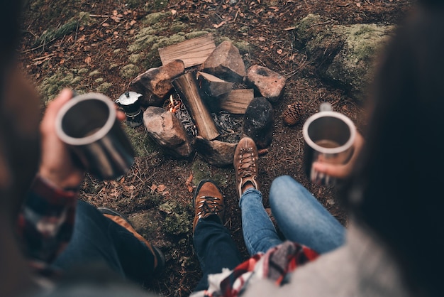 Just relaxing. Close up top view of young couple holding cups while warming up near the campfire