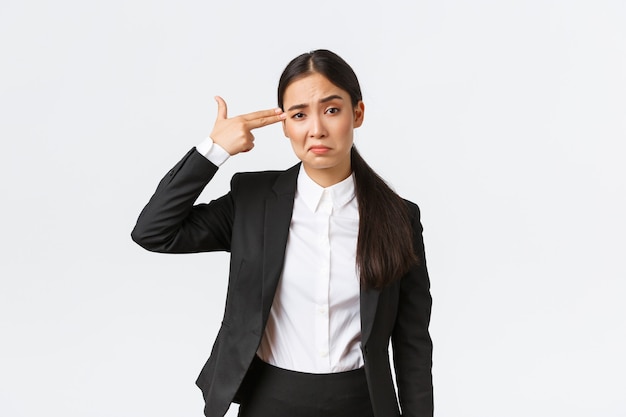 Just shoot me please. Bothered and fed up asian saleswoman hate her work, standing in business suit, shooting herself with hand gun gesture as feeling exhausted, standing white background