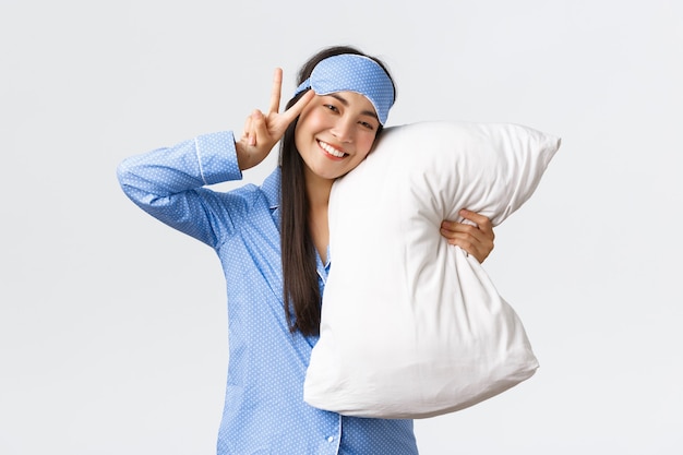 Kawaii happy smiling asian girl in blue sleeping mask and pyjama, hugging pillow and showing peace sign, feeling well after good night sleep, lying in bed during sleepover, white background.