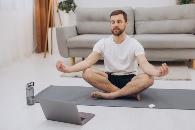 Keep calm on quarantine Millennial guy meditating with trainer online via laptop connection empty space