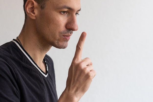 Keep it in secret. Young unshaven man in white tshirt showing silence gesture, making shh and looking at camera with mysterious face expression