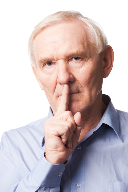 Keep my secret!  Serious senior man in shirt holding finger on lips and looking at camera while standing against white background