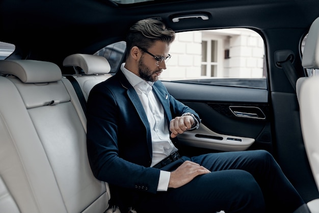 Keeping eye on time. Handsome young man in full suit looking at his watch while sitting in the car