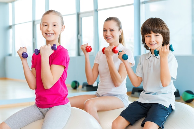 Keeping our bodies fit. Cheerful mother and two children exercising with dumbbells in health club while sitting on the fitness balls together