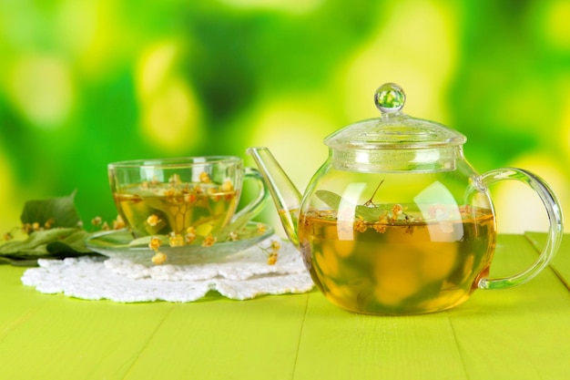 Kettle and cup of tea with linden on wooden table on nature background