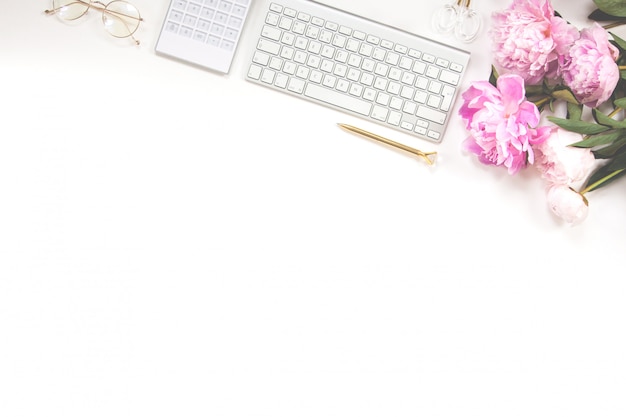 Keyboard, golden pen, glasses, calculator and a bouquet of pink peonies on a white background. Copy space.