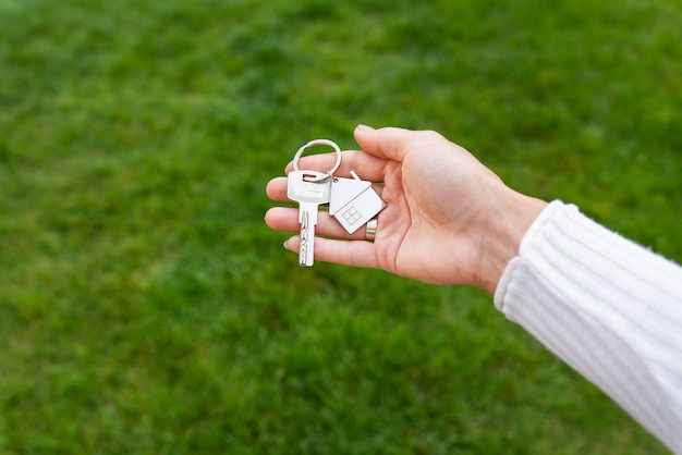 Keys with a keychain in the form of a metal house from a new house or apartment in the hands of a girl against a background of green grass.