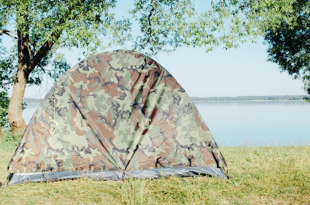 Khaki camping tent on shore against backdrop of lake on sunny summer day Camping in nature hiking concept