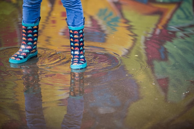 Kid boy in funny rubber boots standing in the puddle in the street after rain