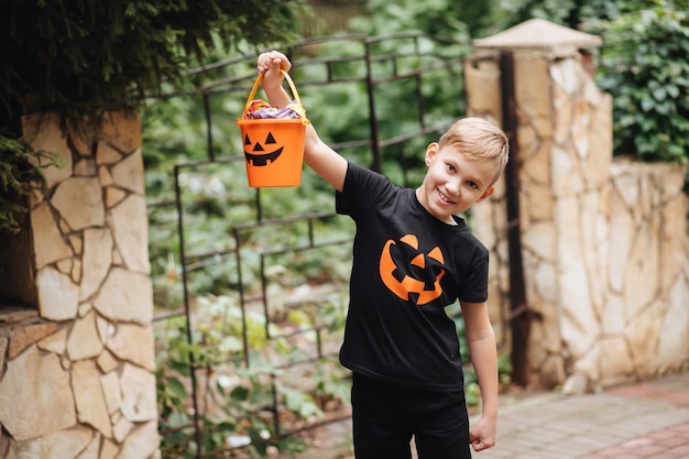 Kid boy teenager holding jack-o-lantern pumpkin bucket with candies and sweets near the fence of the house. Kid trick or treating in Halloween holiday