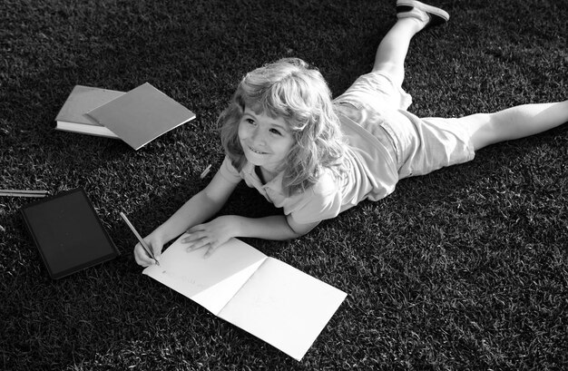 Photo kid boy with pencil writing on notebook lying on grass cute little child in casual clothes reading a book and smiling while lying on grass in park