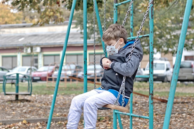 Kid feeling lonely while swinging at  playground and wearing face mask due to coronavirus pandemic