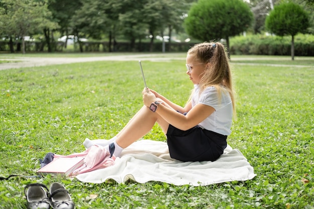 kid girl in glasses and school uniform with wireless headphone has online lesson on tablet on lawn