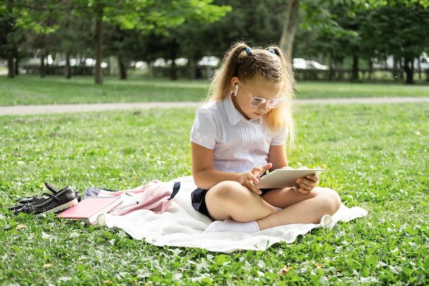 kid girl in glasses and school uniform with wireless headphone has online lesson on tablet on lawn