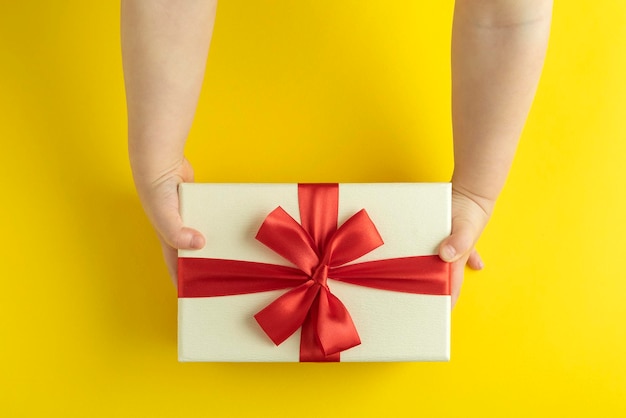 Kid holding gift top view Gift box with red bow in childrens hands on yellow background