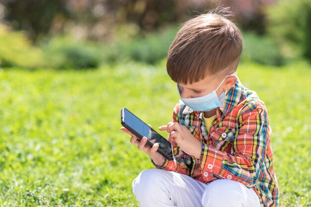 A kid in a medical mask sits on the grass and looks in the phone