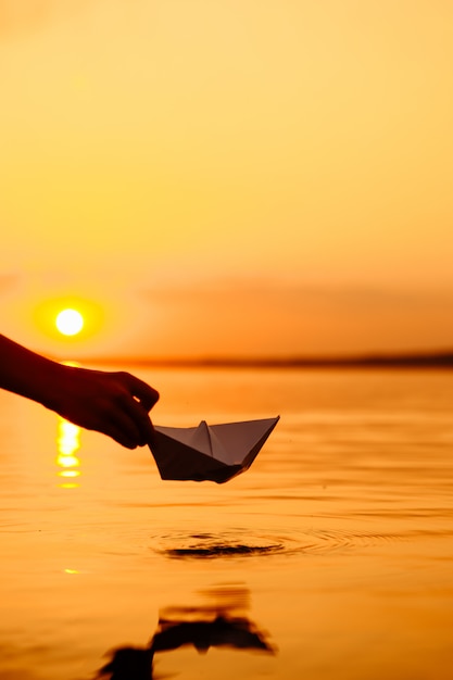 A kid putting a paper boat into water. Beautiful sunset. Origami. River. Lake.