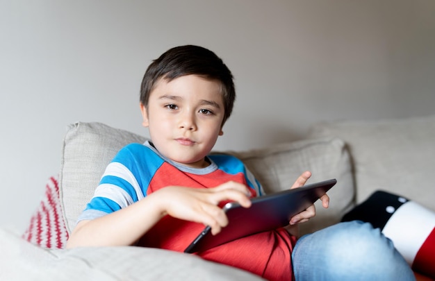 Kid sitting on sofa playing game on tabletYoung Child playing games on internet Portrait School kid using digital pad doing homework online at home Handsome boy looking at camera