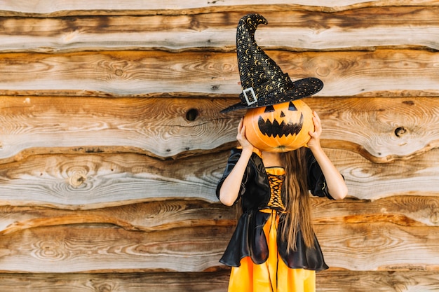 Kid in witch clothes closing face by pumpkin with hat 