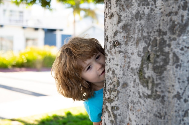 Kids active games. Playing hide and seek. Peekaboo. Little boy hiding by tree.