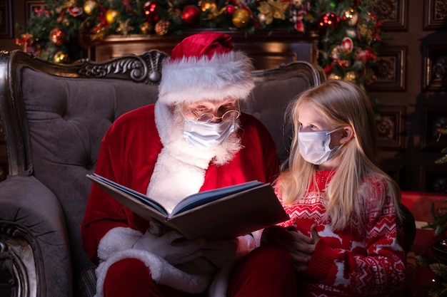 Kids and Santa in medical masks Girl and Santa Claus reading Christmas book sitting near Christmas tree at home Happy New Year Covid 2021