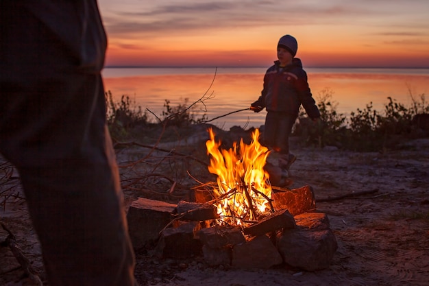 Kids sit near fire on autumn seashore after sunset warm their hands have fun together outside