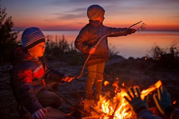 Kids sit near fire on autumn seashore after sunset warm their hands have fun together outside