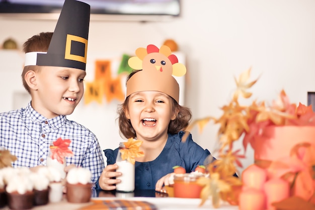 Kids sitting on festive table and celebrating Thanksgiving day. Children in paper turkey hat and pilgrim hats eating cupcakes and drinking milk.