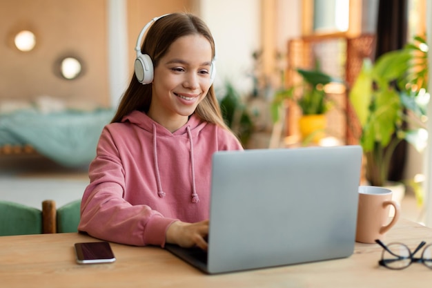 Kids and technology concept Happy teen girl sitting at table and using laptop wearing wireless headphones copy space