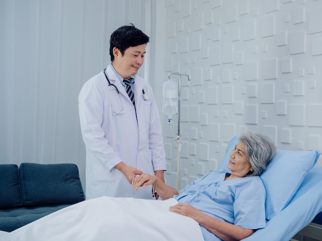 The kind Asian male doctor in white suit visits talks and gives support holding hand of happy elderly senior woman patient in light blue dress lying on bed in saline solution in hospital room